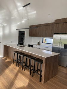 Architectural modern kitchen showing floor-to-ceiling custom woodgrain cabinets, a matching timber island with a white benchtop, and a black linear pendant light, crafted by Hamilton Kitchens