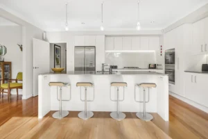 Contemporary custom kitchen design featuring minimalist white cabinetry, a large central island bench with chrome bar stools, and polished timber floors by Hamilton Kitchens