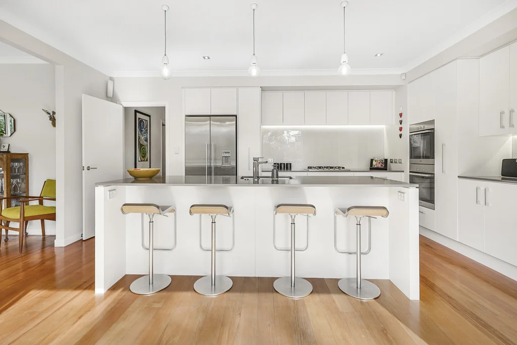 Contemporary custom kitchen design featuring minimalist white cabinetry, a large central island bench with chrome bar stools, and polished timber floors by Hamilton Kitchens