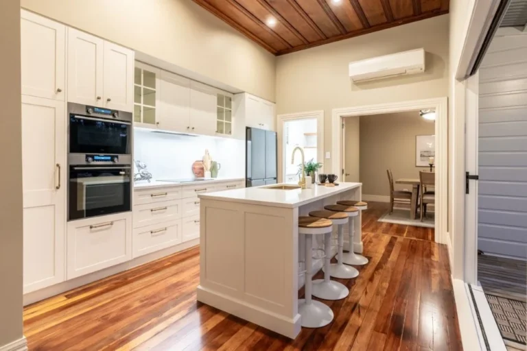High-end kitchen renovation featuring a custom wall tower with integrated double ovens, white shaker pantry cabinetry, a central island with a gold tap, and rich timber floors by Hamilton Kitchens