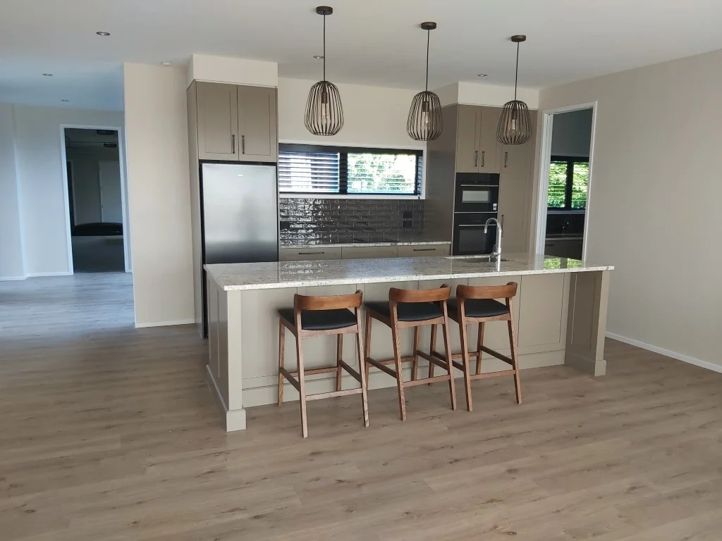 Transitional kitchen design featuring taupe shaker-style cabinets, a central island with a granite benchtop, three timber bar stools, and black wire pendant lights by Hamilton Kitchens